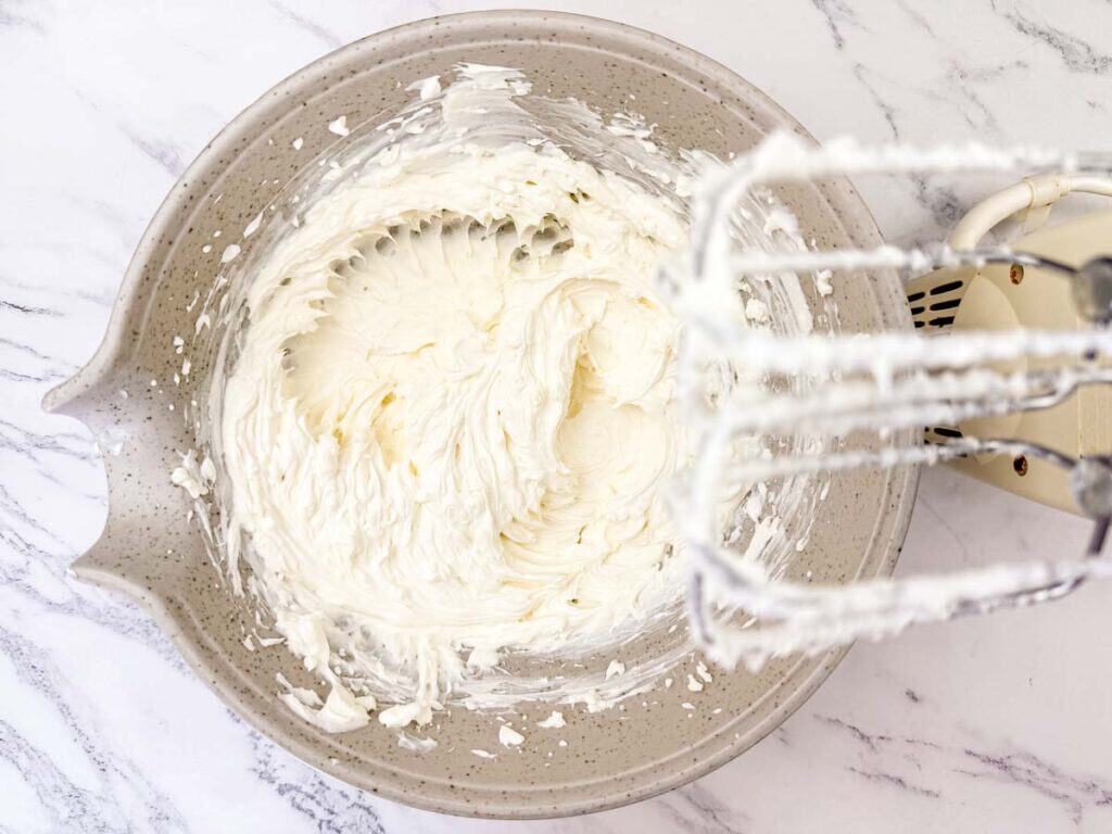 A bowl of whipped cream cheese mixture with an electric hand mixer and beaters resting on the side, viewed from above on a marble surface.