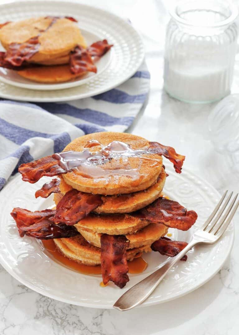Stack of pancakes with crispy bacon strips, syrup, and butter on a white plate with a fork; second plate and striped towel in the background.