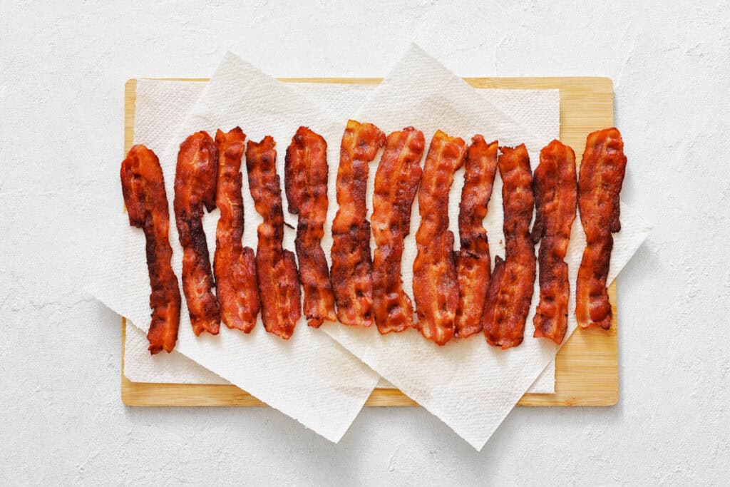 Crispy cooked bacon strips arranged in a row on paper towels atop a wooden cutting board.