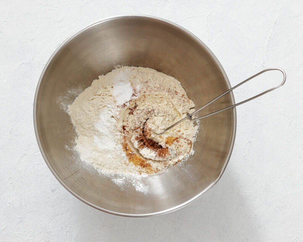 A metal mixing bowl with flour, baking powder, and spices, partially mixed with a metal whisk, on a white surface.