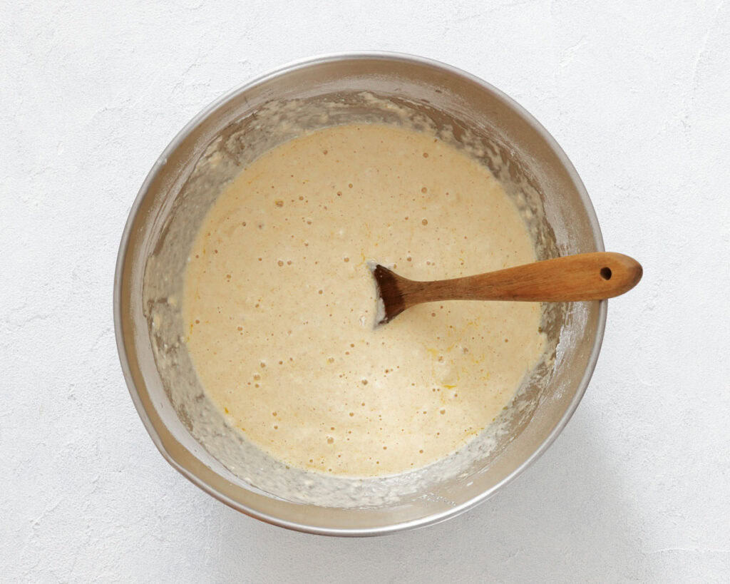 A metal mixing bowl filled with pancake batter and a wooden spoon resting inside, placed on a light-colored surface.