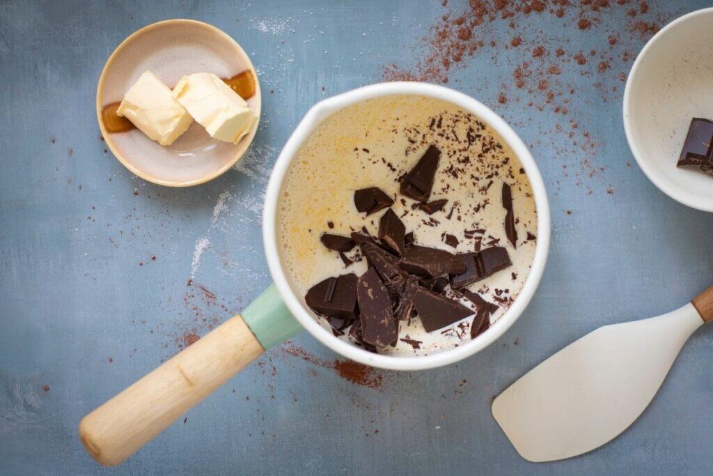 A saucepan filled with cream and dark chocolate pieces sits on a blue surface, next to two butter cubes on a plate, a spatula, and an empty bowl.