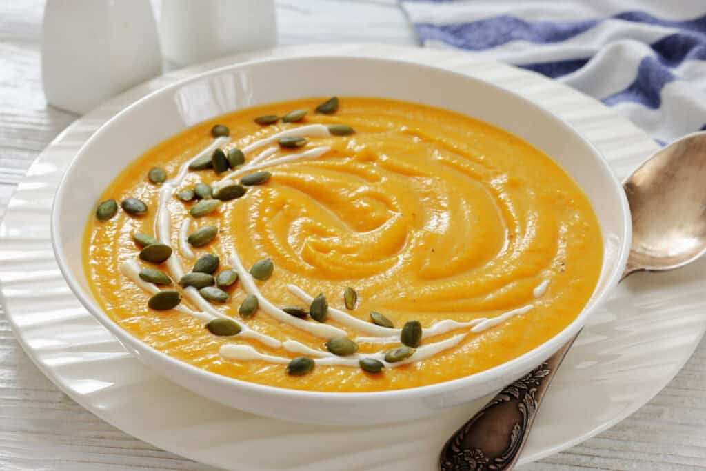 A bowl of creamy orange soup garnished with a swirl of cream and sprinkled with pumpkin seeds, with a spoon on the side.