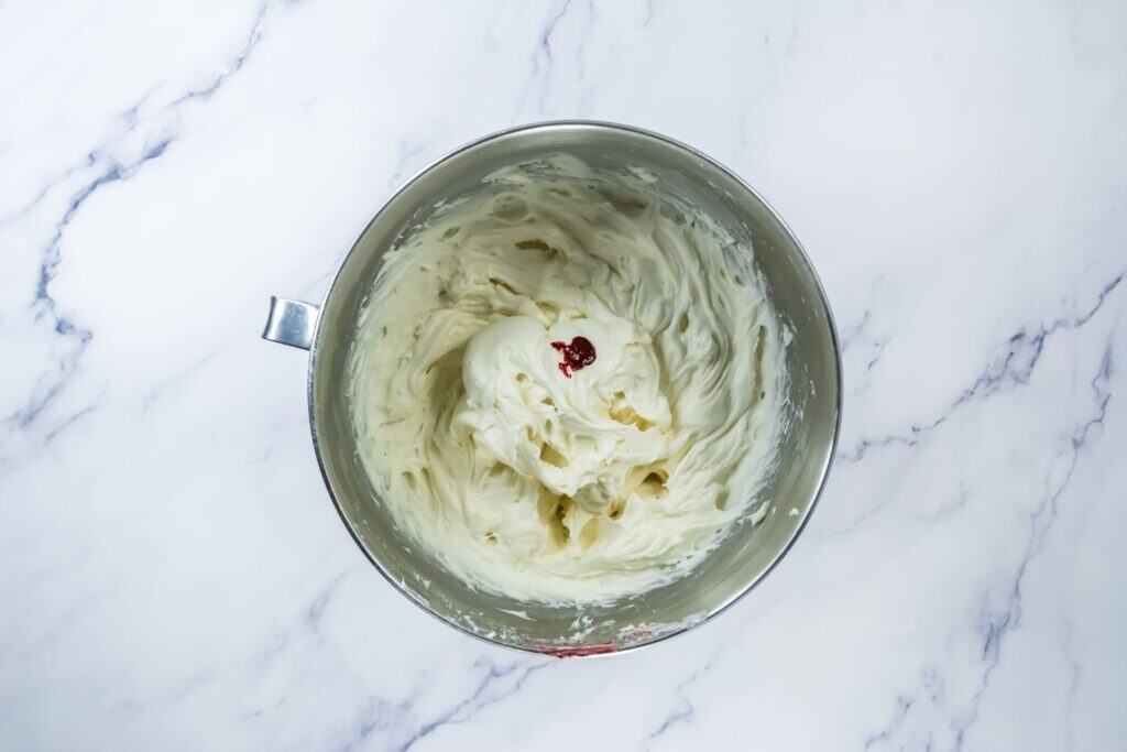 A metal mixing bowl containing thick white frosting with a small drop of red food coloring in the center, placed on a white marble surface.