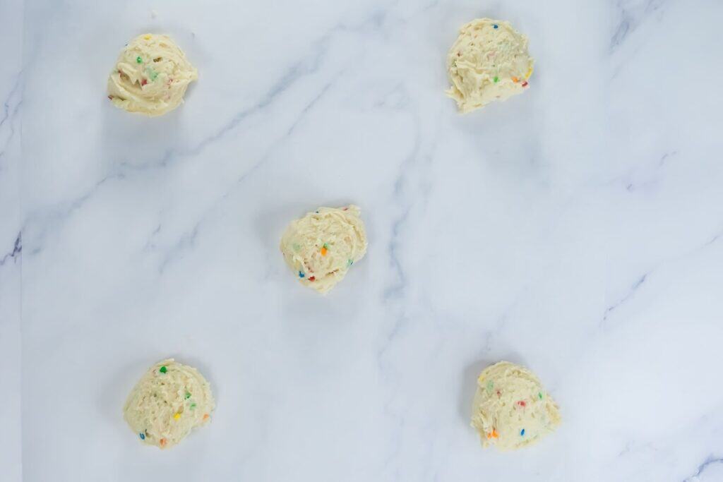 Five scoops of raw cookie dough with colorful sprinkles are spaced out on a white marble surface.