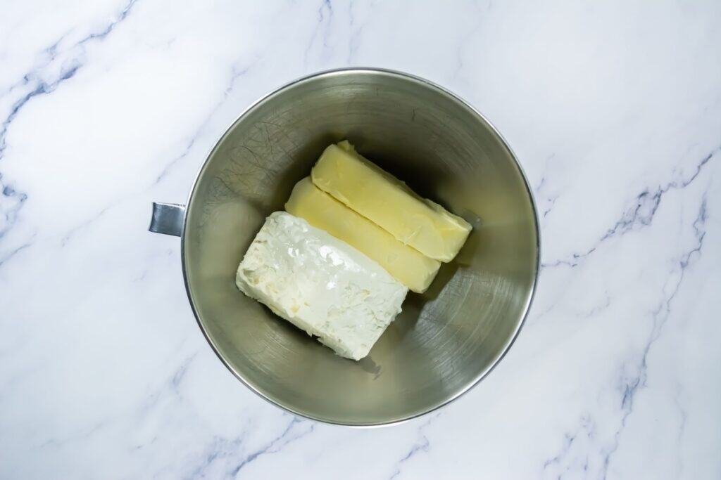 A metal mixing bowl containing two sticks of butter and a block of cream cheese on a white marble surface.