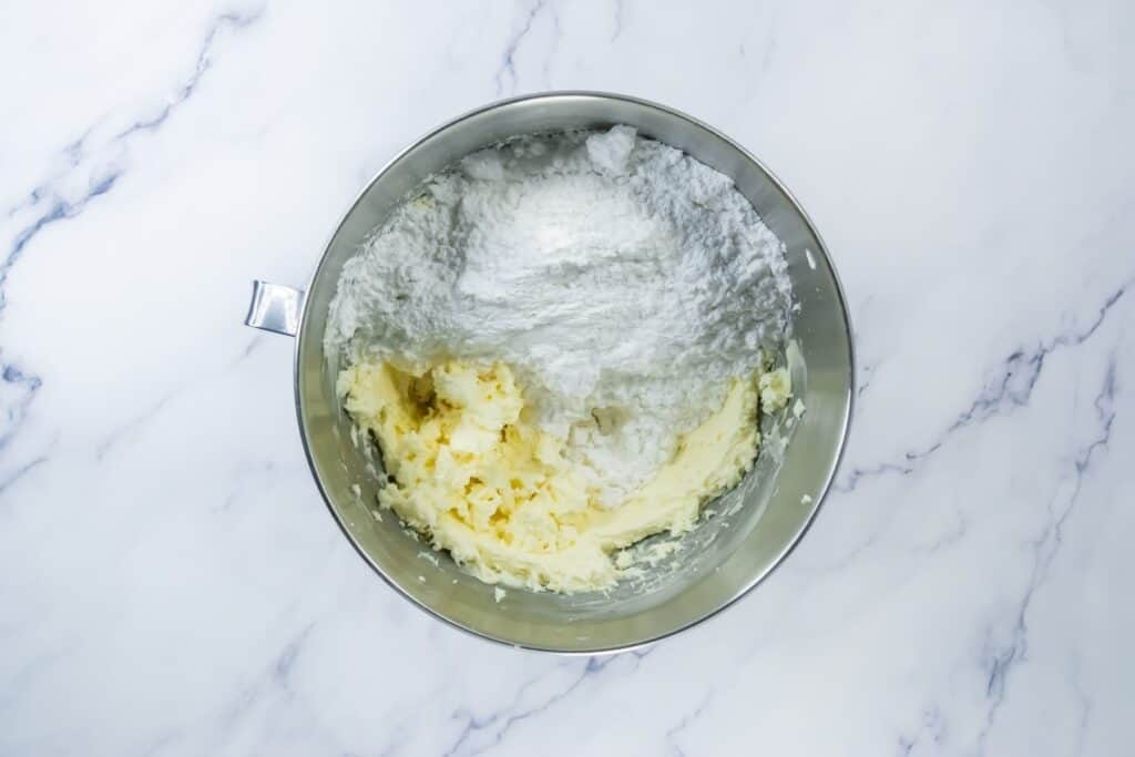 A metal mixing bowl containing softened butter and powdered sugar on a white marble surface, ready to be mixed.