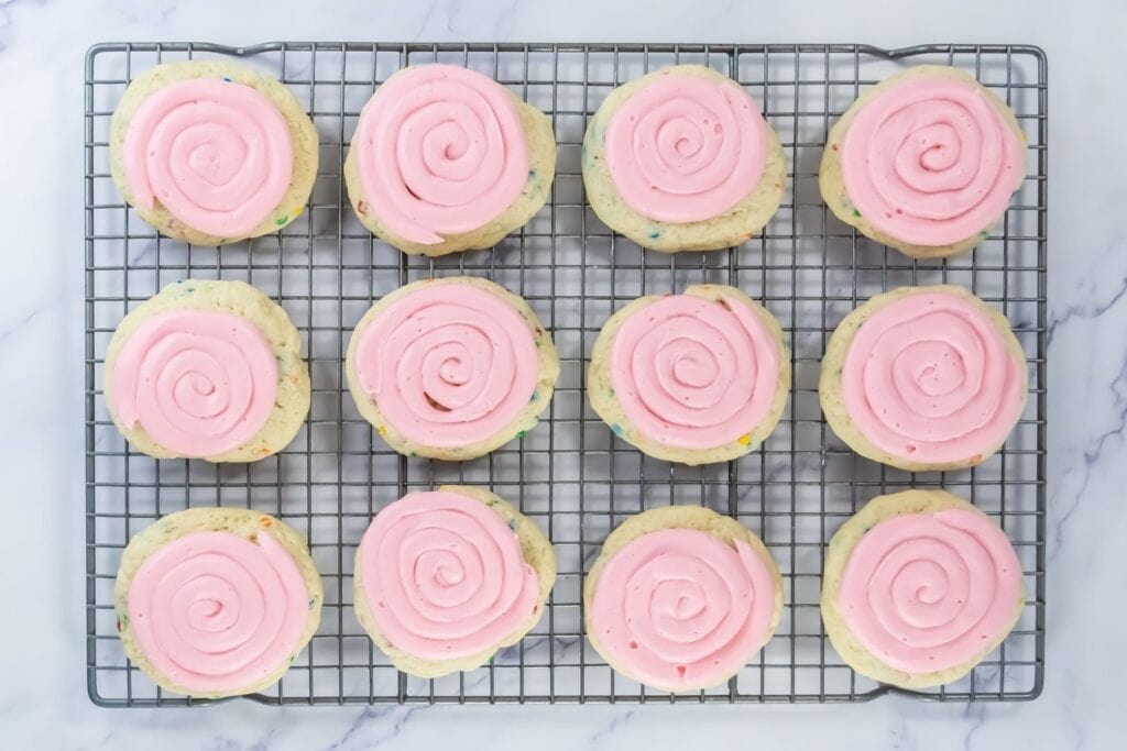 Twelve cookies with pink swirl frosting are arranged in rows on a wire cooling rack over a marble surface.