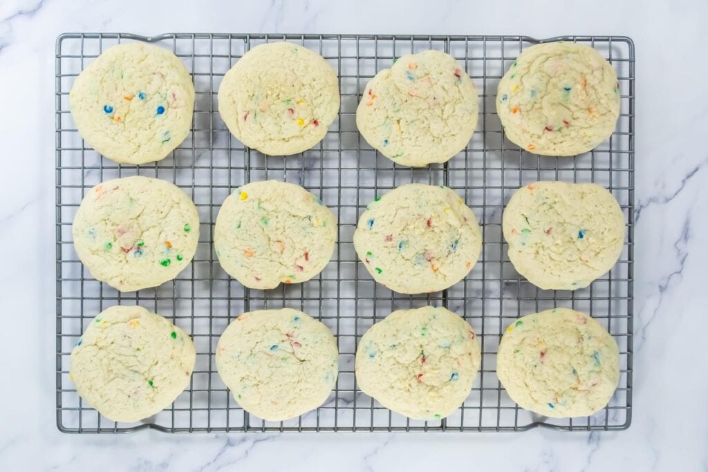 Twelve round cookies with colorful sprinkles are arranged in rows on a cooling rack over a white marble surface.