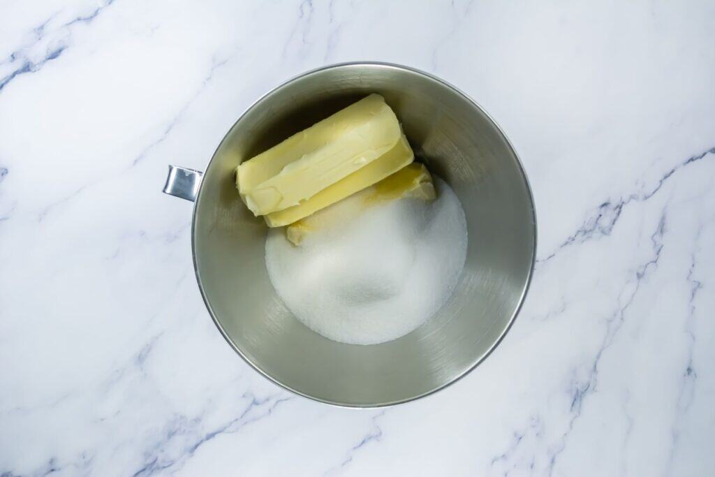 A metal mixing bowl containing two sticks of butter and a mound of granulated sugar, placed on a white marble surface.