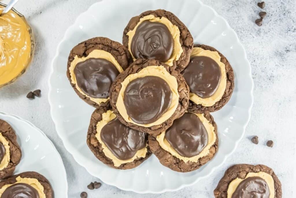 A white plate with eight chocolate cookies topped with a layer of peanut butter and a layer of chocolate frosting. Scattered chocolate chips and a jar of peanut butter are nearby.