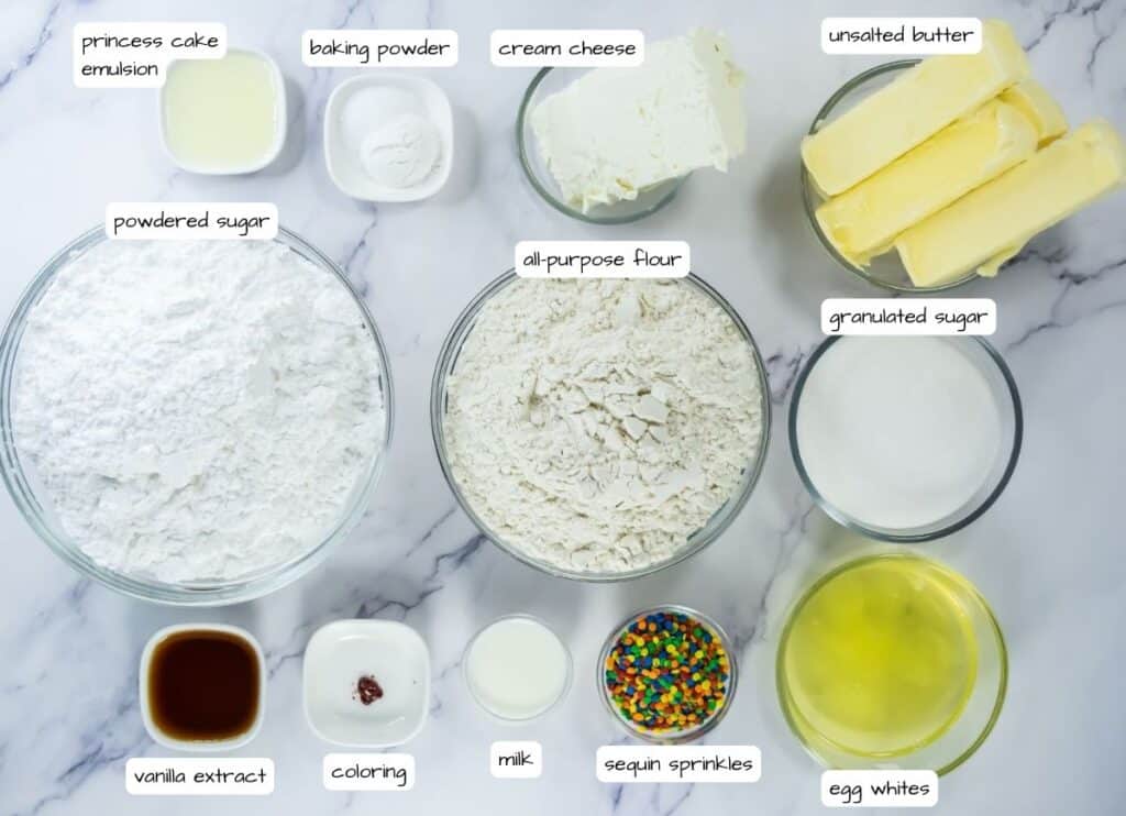 Top-down view of various baking ingredients in bowls on a marble surface, including flour, sugar, butter, cream cheese, egg whites, milk, and labeled flavorings and decorations.