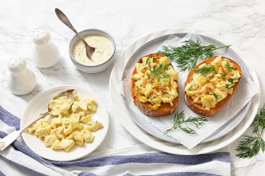 Two pieces of toast topped with egg salad and fresh dill on a plate, with extra egg salad on a side plate and a bowl of sauce nearby. Salt and pepper shakers in the background.