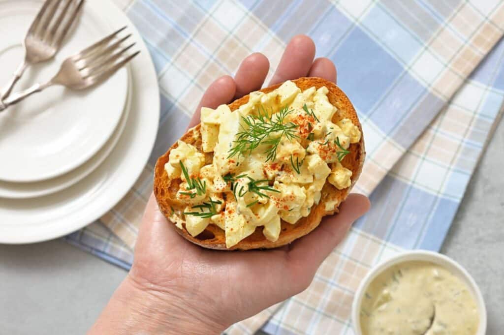A hand holds a slice of toast topped with egg salad and fresh dill, with a stack of plates, forks, and a bowl of dip on a plaid tablecloth in the background.