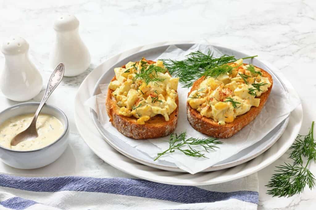 Two slices of bread topped with egg salad and fresh dill on a white plate, with a bowl of sauce, a spoon, salt and pepper shakers, and a blue-striped napkin nearby.