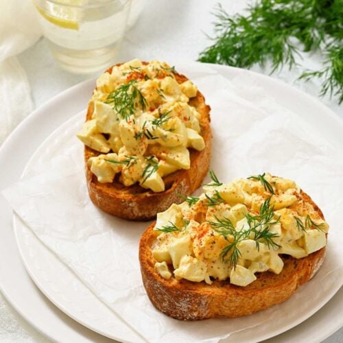 Two slices of toasted bread topped with chopped egg salad and fresh dill on a white plate, with a glass of water and sprigs of dill nearby.