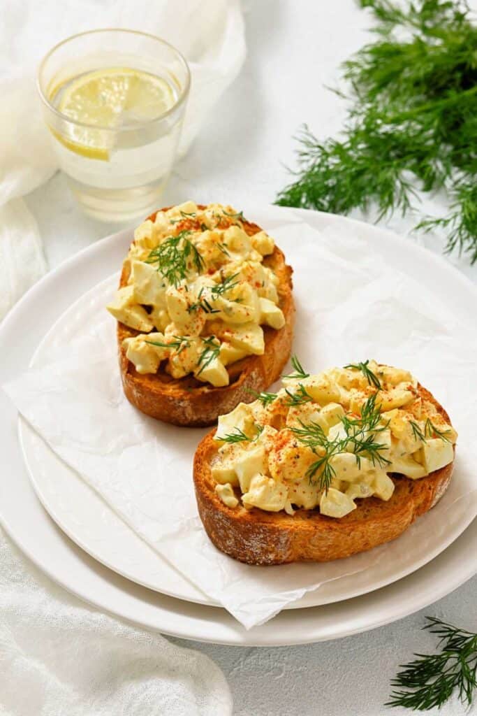 Two slices of toasted bread topped with chopped egg salad and fresh dill on a white plate, with a glass of water and sprigs of dill nearby.