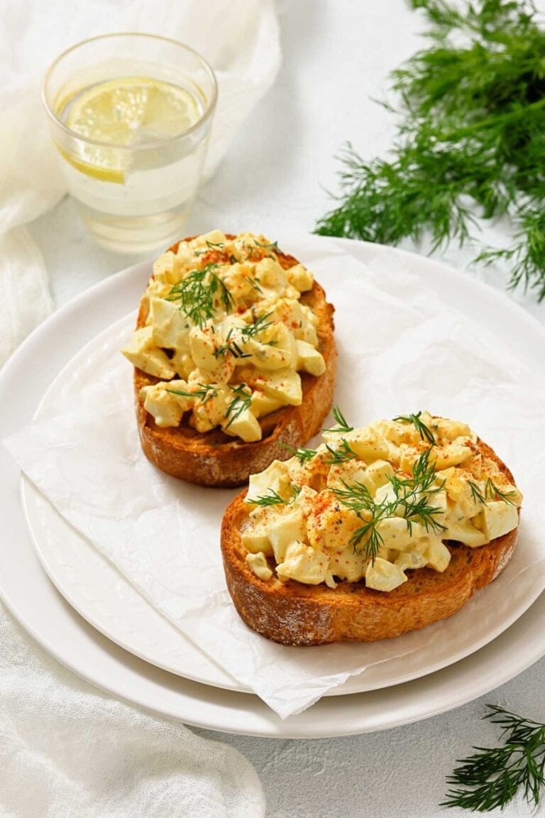 Two slices of toasted bread topped with chopped egg salad and fresh dill on a white plate, with a glass of water and sprigs of dill nearby.