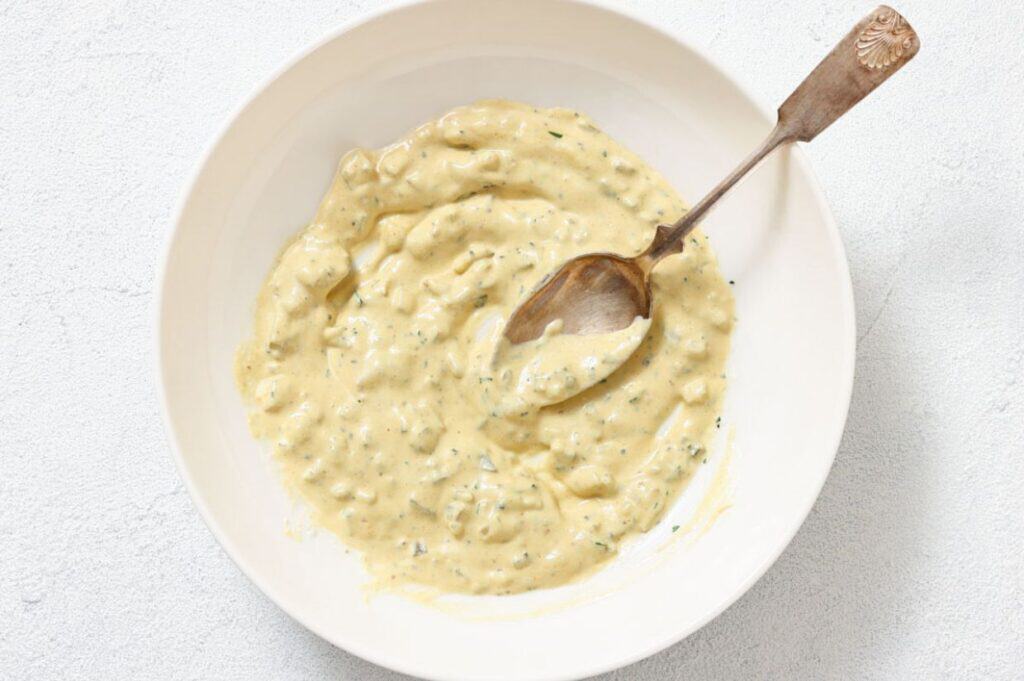 A white bowl filled with a thick, creamy sauce mixed with herbs and small chunks, with a metal spoon resting inside.