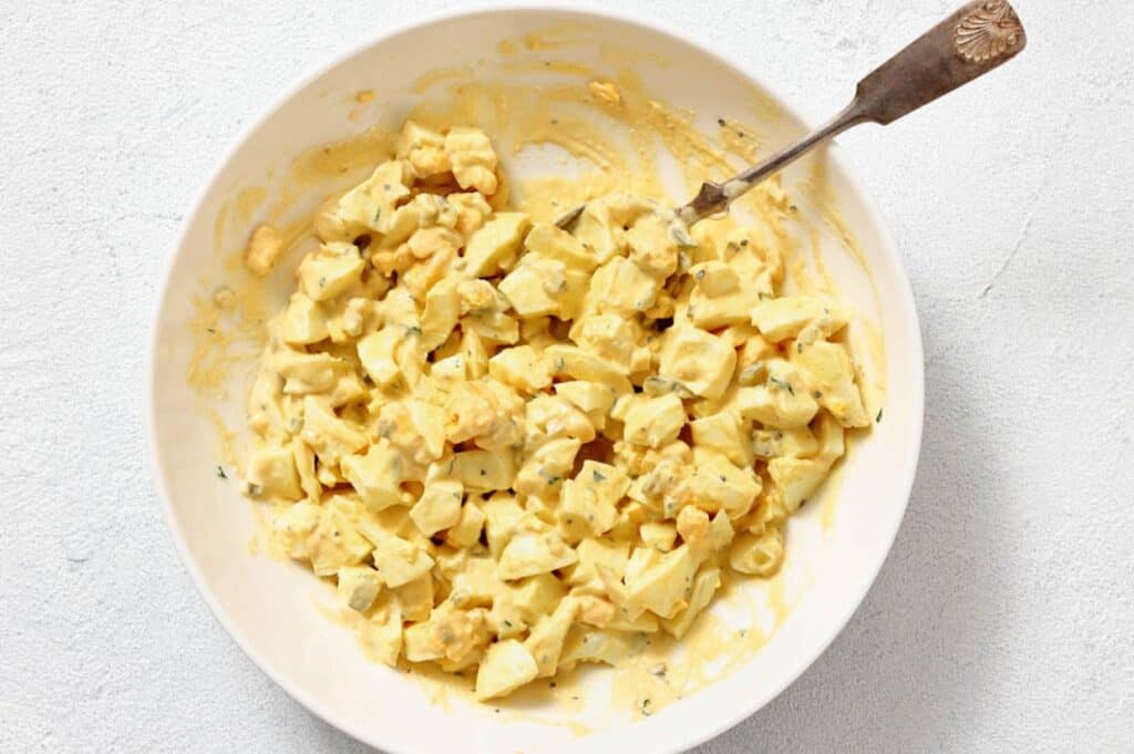 A bowl of chopped egg salad mixed with mayonnaise and seasonings, with a spoon resting in the bowl.