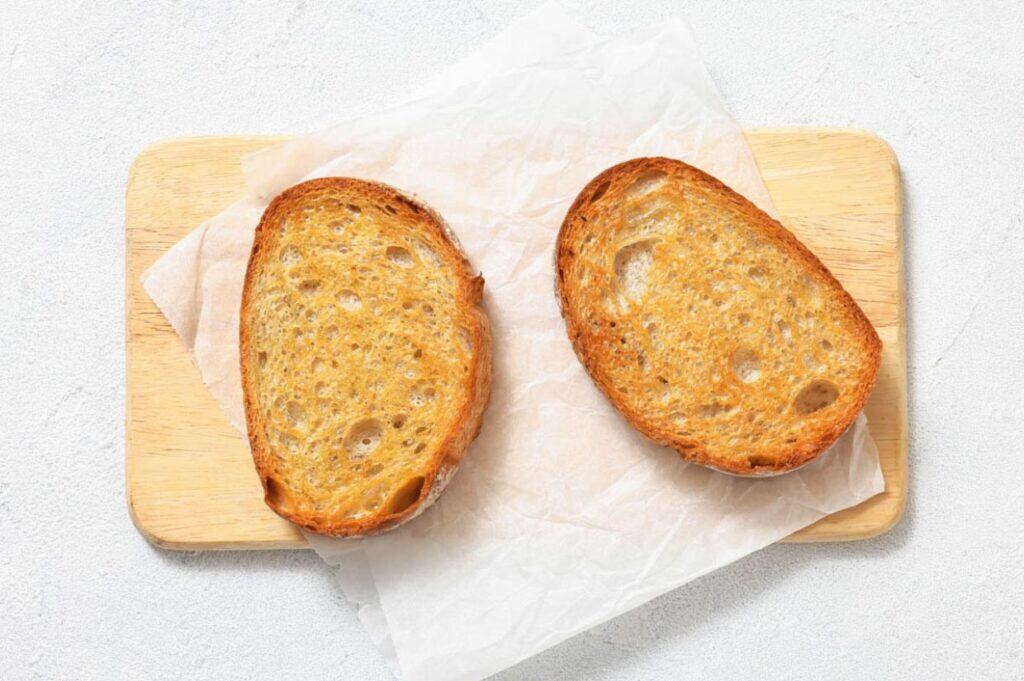Two slices of toasted bread on a sheet of parchment paper atop a wooden cutting board.
