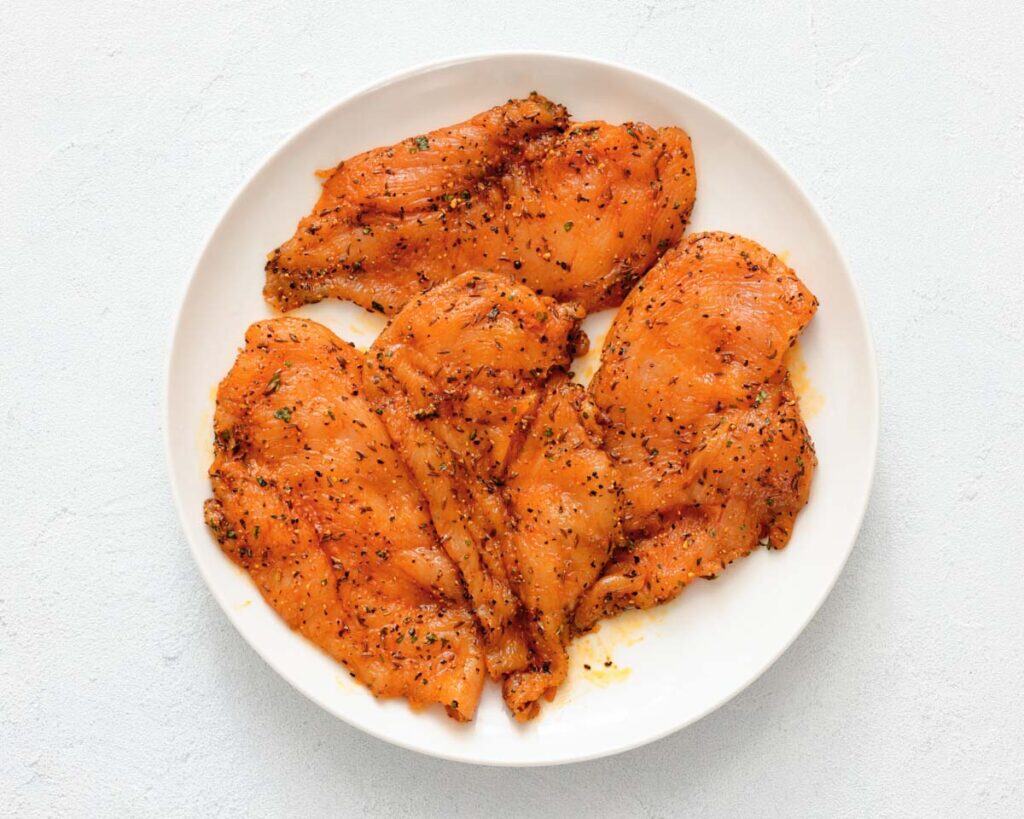 Four seasoned raw chicken breasts arranged on a white plate against a light background.