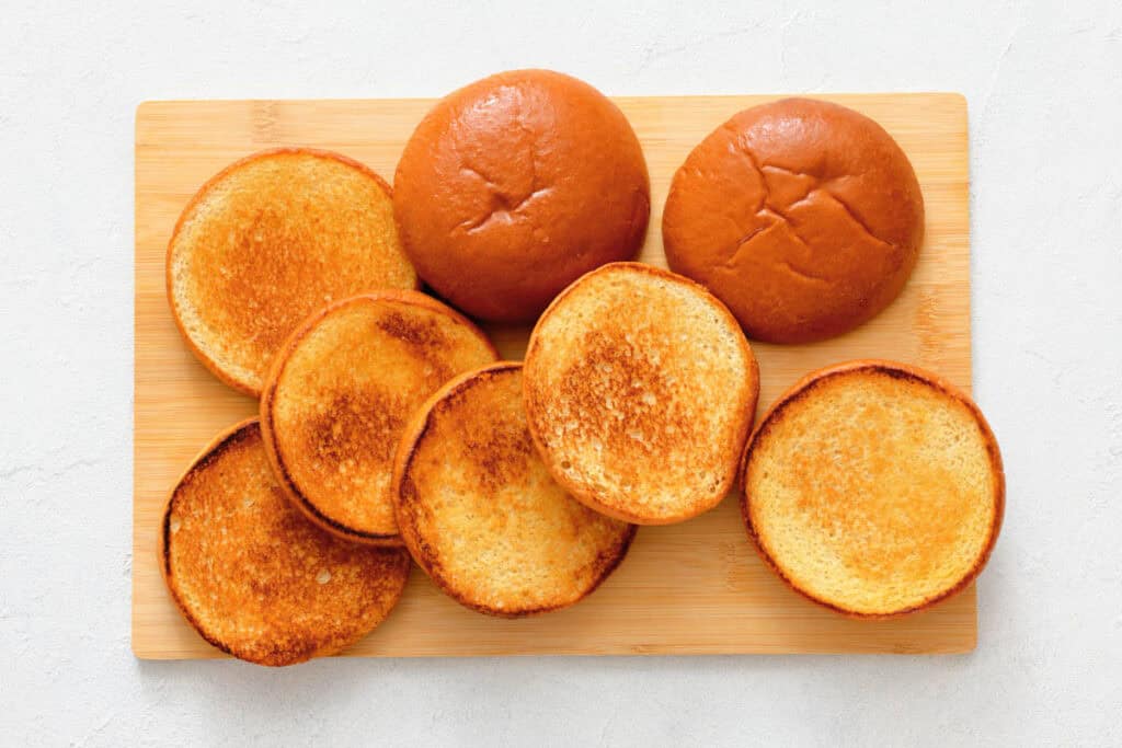 A wooden board with two whole sandwich buns and six toasted sandwich bun halves arranged on top.