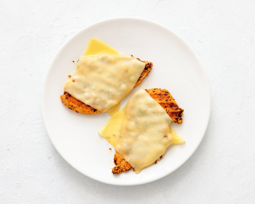 Two pieces of cooked chicken breast topped with melted cheese slices on a white plate.