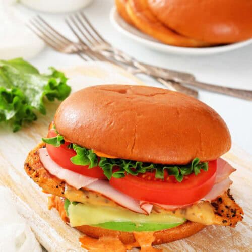 A chicken sandwich with lettuce, tomato, ham, cheese, and sauce on a brioche bun, served on a wooden board with a plate of buns and sauce in the background.