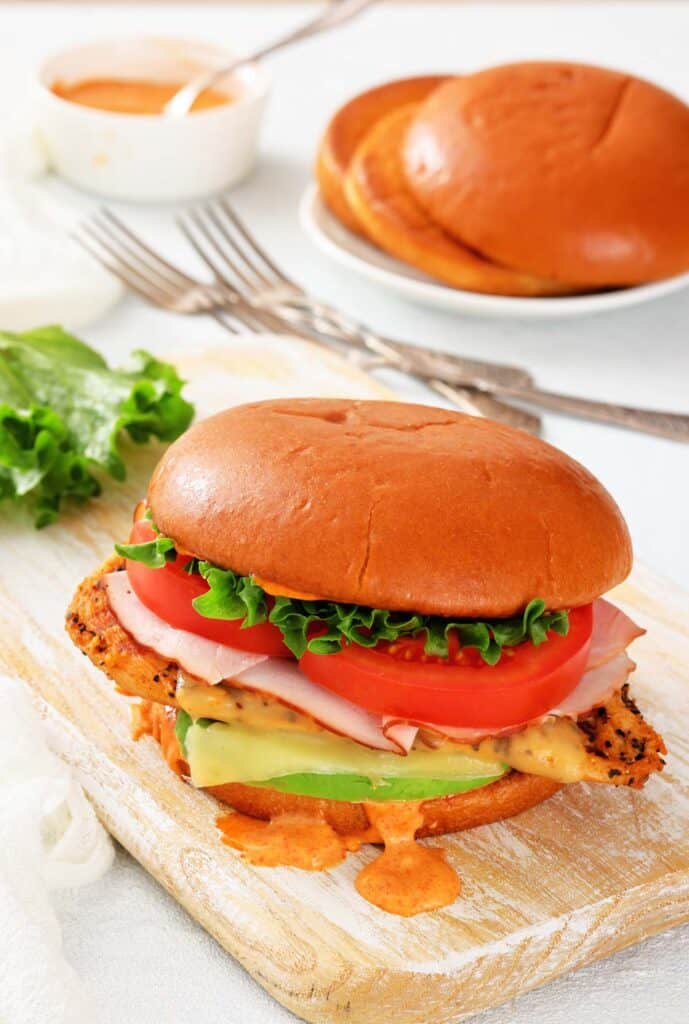 A chicken sandwich with lettuce, tomato, ham, cheese, and sauce on a brioche bun, served on a wooden board with a plate of buns and sauce in the background.