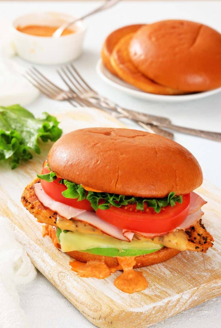 A chicken sandwich with lettuce, tomato, ham, cheese, and sauce on a brioche bun, served on a wooden board with a plate of buns and sauce in the background.