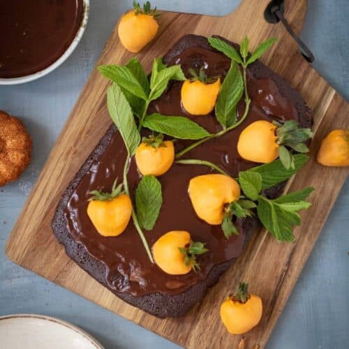 A chocolate cake with glossy icing is topped with yellow candy-coated treats and green mint leaves, displayed on a wooden board with a bowl of chocolate, utensils, and a gold spider nearby.