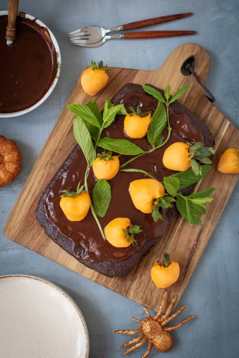 A chocolate cake with glossy icing is topped with yellow candy-coated treats and green mint leaves, displayed on a wooden board with a bowl of chocolate, utensils, and a gold spider nearby.