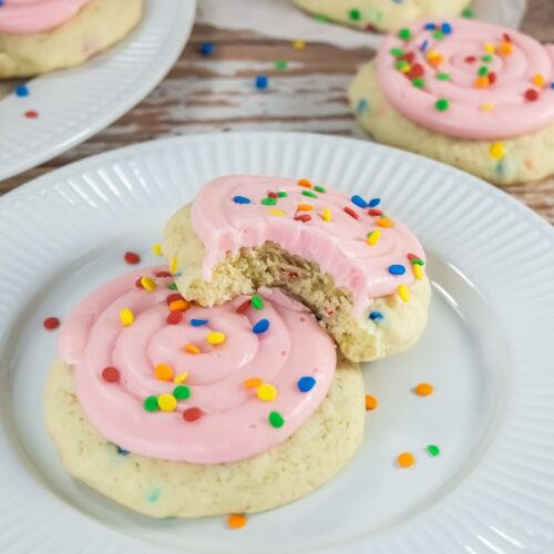 Two sugar cookies with pink frosting and colorful sprinkles on a white plate; one cookie has a bite taken out. More cookies are stacked in the background.