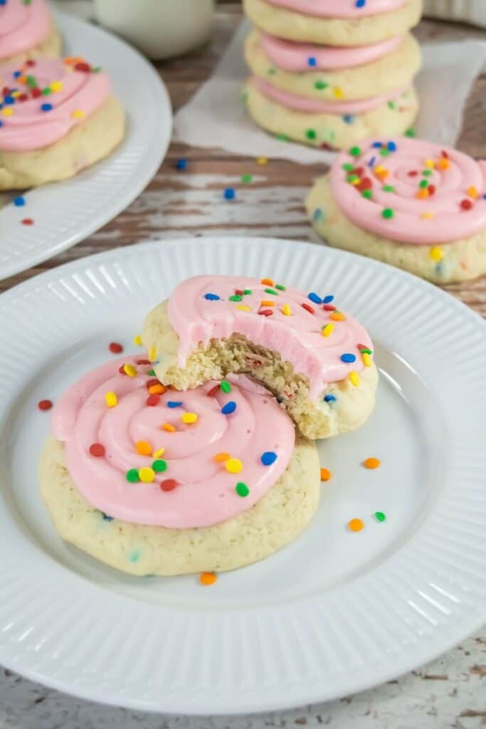 Two sugar cookies with pink frosting and colorful sprinkles on a white plate; one cookie has a bite taken out. More cookies are stacked in the background.