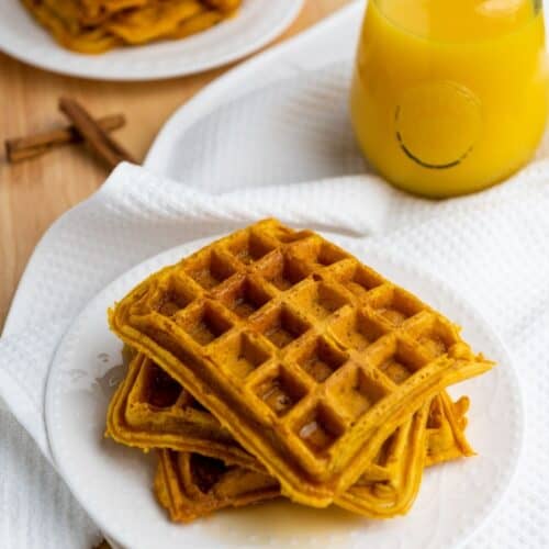 A plate of stacked Pumpkin Waffles sits on a white cloth next to a glass of juice, with more waffles on a plate in the background.