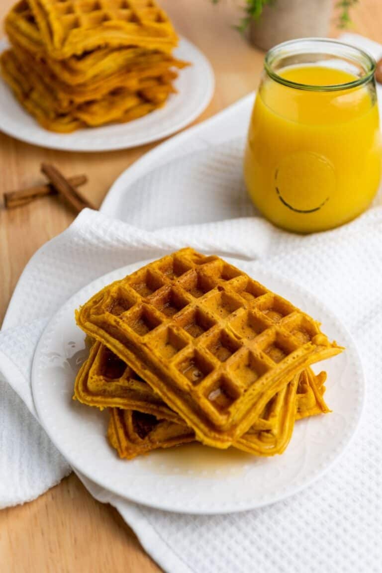 A plate of stacked Pumpkin Waffles sits on a white cloth next to a glass of juice, with more waffles on a plate in the background.
