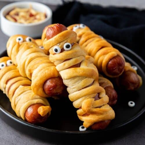 A plate of hot dogs wrapped in dough to look like mummies, with candy eyes added for decoration; a small bowl of nuts is in the background.