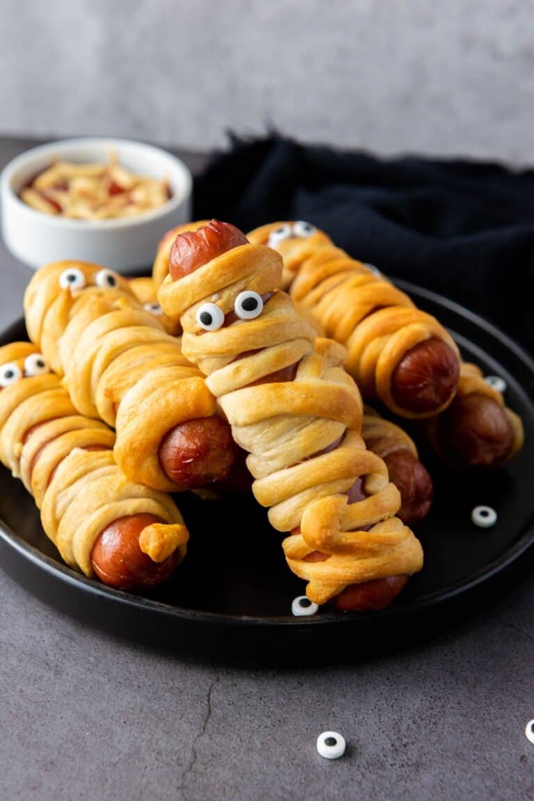 A plate of hot dogs wrapped in dough to look like mummies, with candy eyes added for decoration; a small bowl of nuts is in the background.