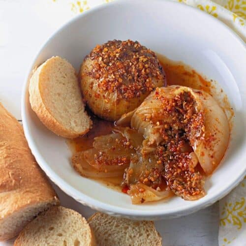 A white bowl with baked onions topped with spices and sauce, served with slices of baguette bread on a white surface.