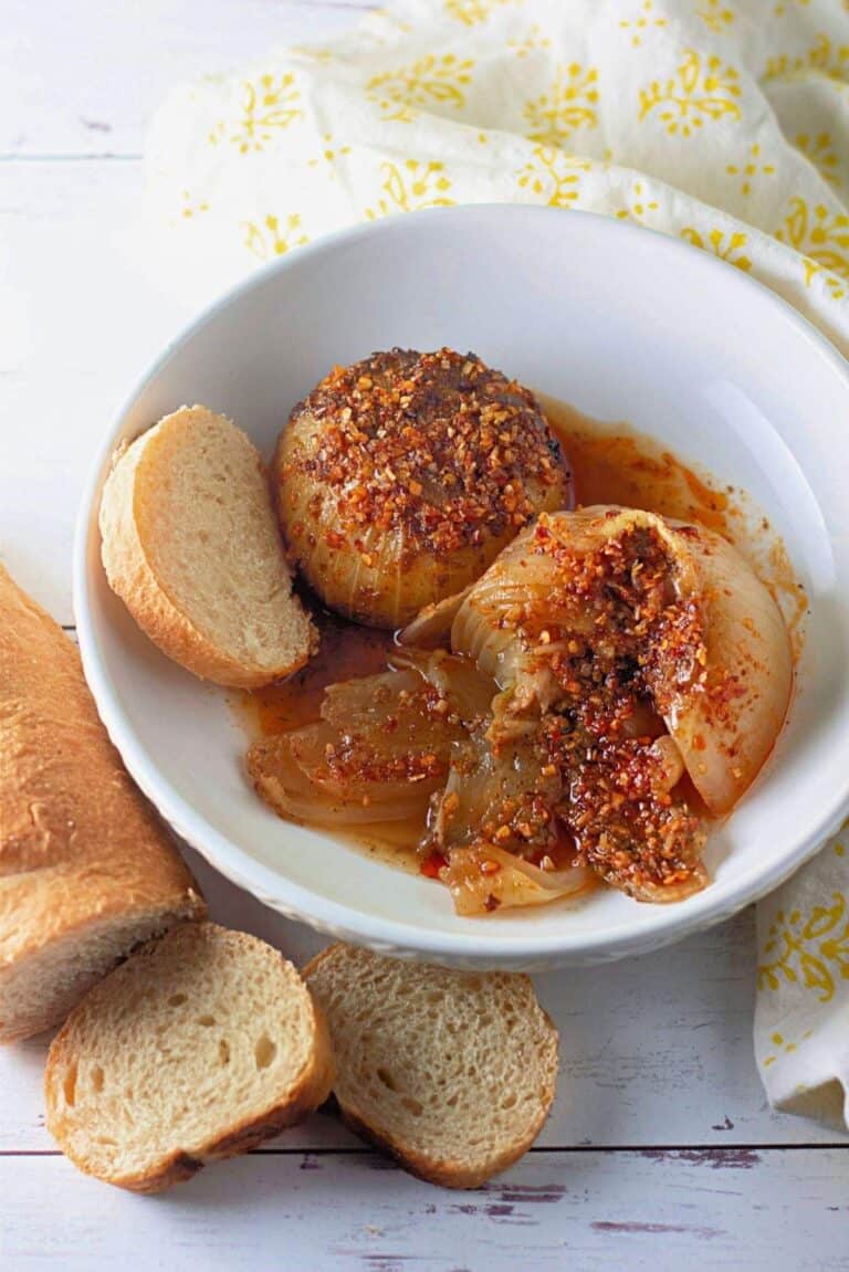 A white bowl with baked onions topped with spices and sauce, served with slices of baguette bread on a white surface.