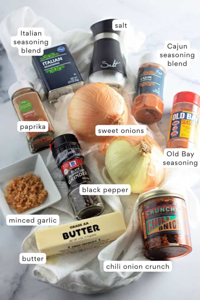 Various seasonings and ingredients, including Italian seasoning, Cajun seasoning, salt, paprika, Old Bay, sweet onions, garlic, black pepper, butter, and chili onion crunch, on a white surface.