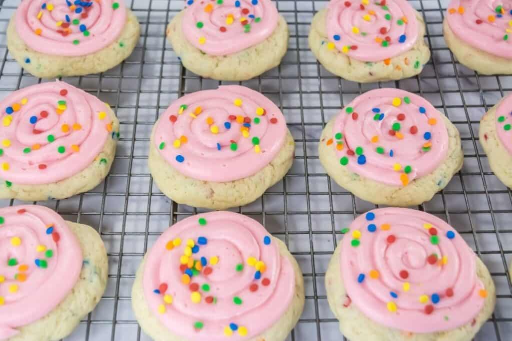 Sugar cookies with pink frosting and colorful round sprinkles are arranged on a cooling rack in rows.