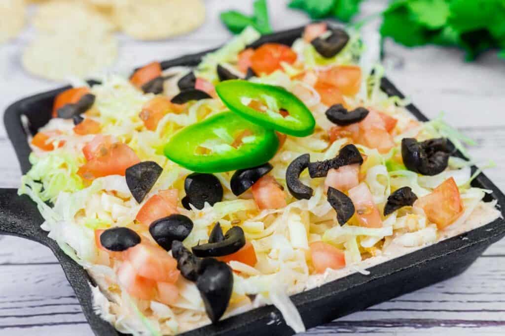 A square dish filled with layered dip topped with shredded lettuce, diced tomatoes, sliced black olives, and jalapeño slices.