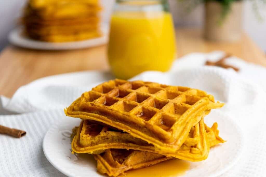 A stack of waffles on a white plate with syrup, with a glass of orange juice and more waffles in the background.
