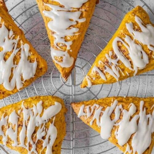 Five Pumpkin Spice scones with vanilla glaze are arranged in a circle on a round metal cooling rack over a light-colored surface.