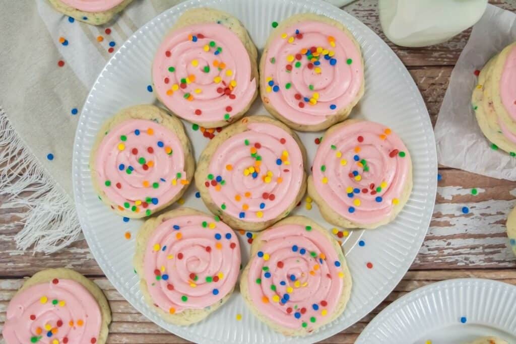 A plate of round sugar cookies topped with pink frosting and multicolored sprinkles, arranged on a white plate on a wooden surface.
