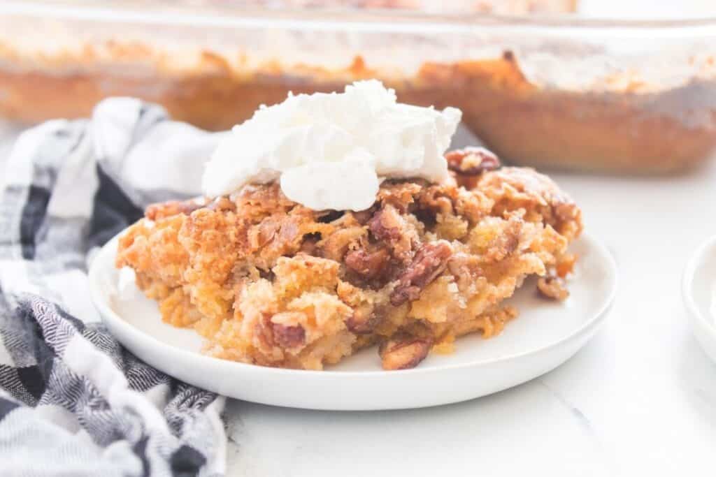 A serving of pecan-topped dessert with a dollop of whipped cream on a white plate, with a checkered cloth nearby.