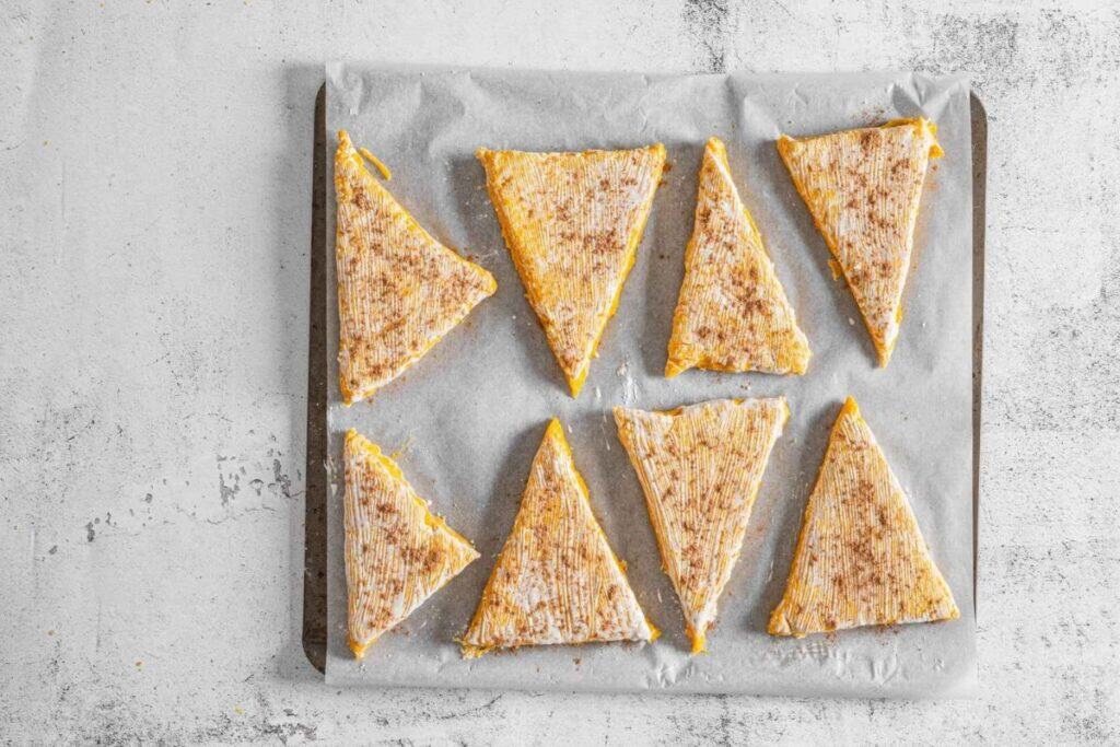 Eight triangular scones dusted with pumpkin pie spice and coated with cream are arranged on a parchment-lined baking sheet.