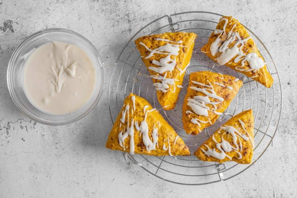 Five triangular pastries with white glaze drizzle on a cooling rack next to a bowl of glaze, all on a light textured surface.